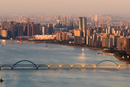 Hangzhou Qiantang River City Skyline at Golden Hour View of Qiantang River with arch railway bridge, dense modern skyscrapers, Hangzhou Olympic Sports Center and calm river surface under warm sunset glow