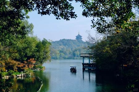 Peaceful view of West Lake in Hangzhou with a traditional boat, green trees, and Leifeng Pagoda in the distance