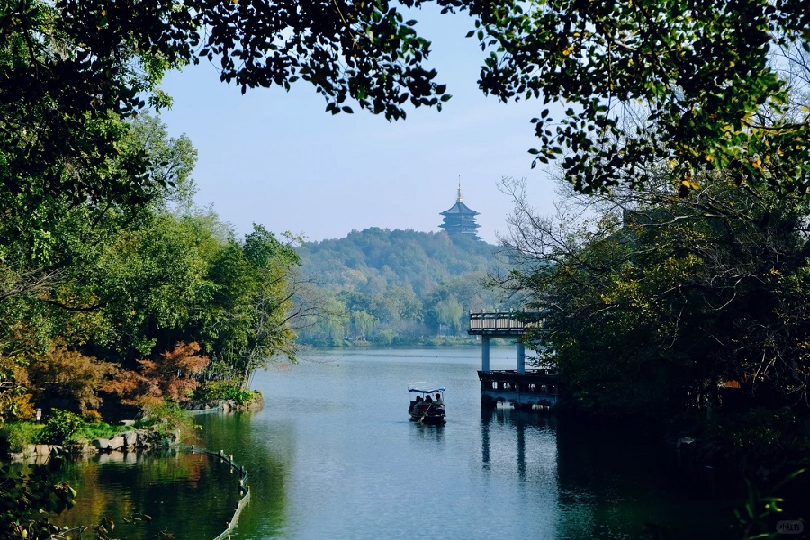 Peaceful view of West Lake in Hangzhou with a traditional boat, green trees, and Leifeng Pagoda in the distance