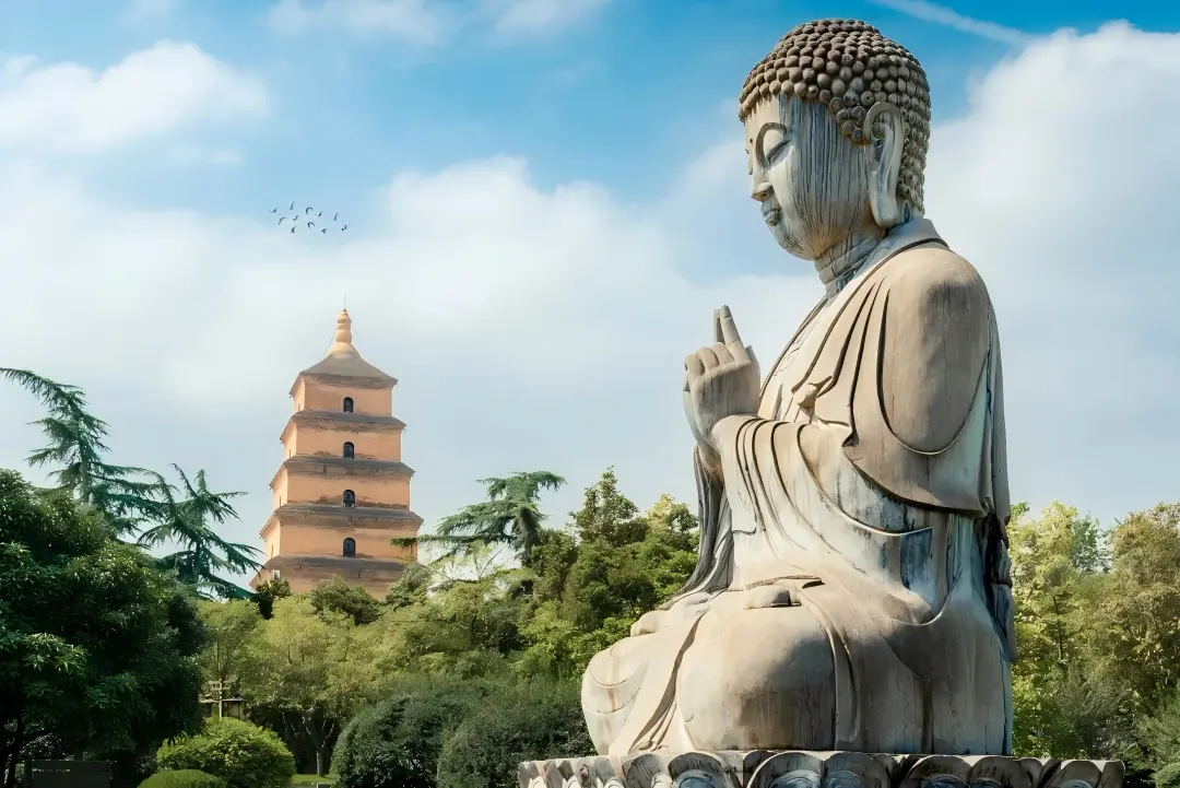 Stone Buddha statue in foreground, Giant Wild Goose Pagoda surrounded by green trees under blue cloudy sky