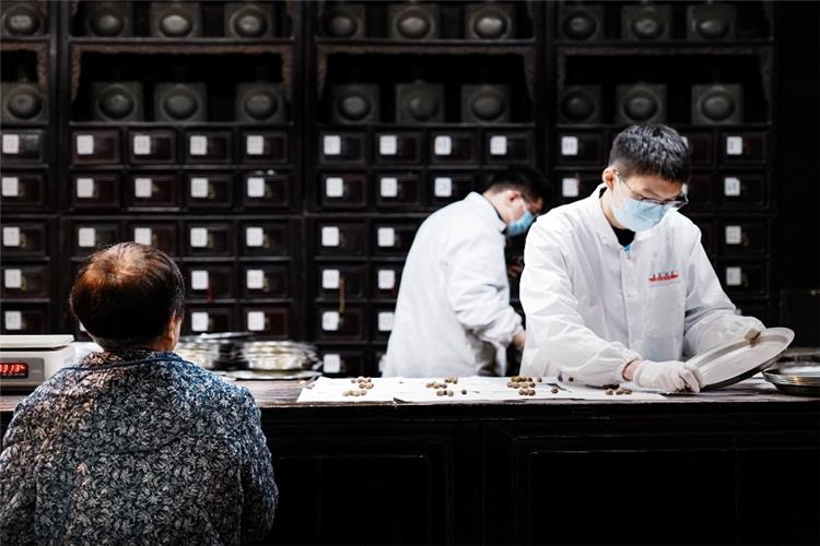 Professional TCM pharmacists in white lab coats weighing medicinal herbs for customers in front of ancient wooden medicine cabinets inside Hu Qing Yu Tang pharmacy