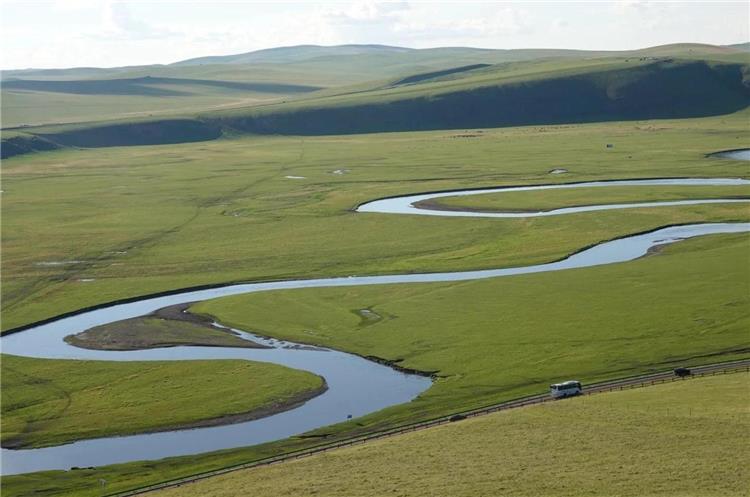 Aerial view of meandering Morigele River flowing across vast green Inner Mongolia grassland, road with tourist vehicles