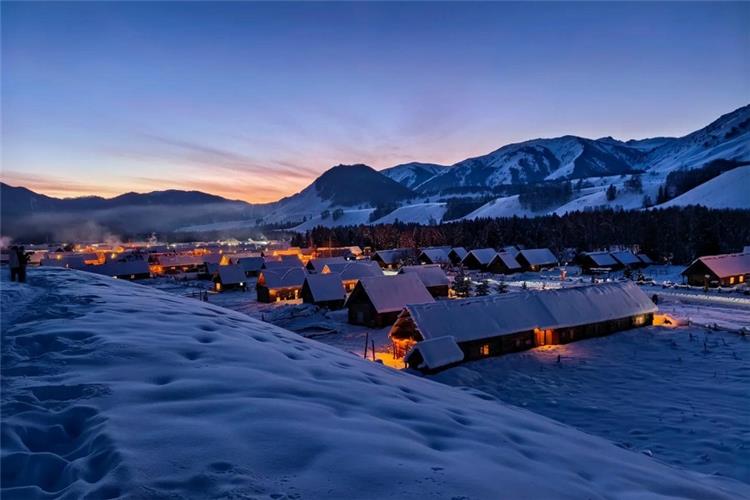 Magical winter dusk scenery of snow?blanketed Hemu village, warm lights inside wooden houses against snow mountain range
