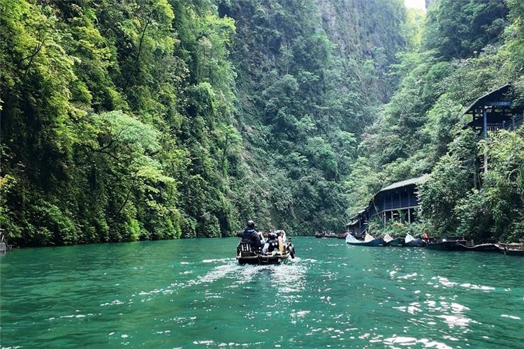 Tourist boat sailing on turquoise river in Pingshan Canyon, steep green cliffs and wooden stilt houses