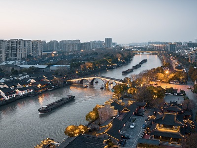 Stunning aerial dusk scenery of Gongchen Stone Arch Bridge along the ancient Grand Canal, featuring traditional Chinese architecture, river cargo boats and illuminated ancient streets in Hangzhou
