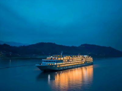 Illuminated luxury cruise vessel sailing on calm Yangtze River, dark mountain backdrop at blue hour dusk