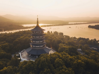 Aerial shot of multi-tiered Leifeng Pagoda surrounded by dense woods, golden sunset glow reflecting on West Lake, classic Hangzhou landmark