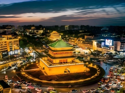 Overhead shot of illuminated Xi'an Bell Tower with green glazed roof, busy traffic roundabout, lit Drum Tower in background, sunset gradient sky blending ancient architecture and modern city night sce