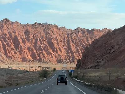 Scenic road through Xinjiang canyon with red rock mountains, desert landscape, and vehicles traveling under a blue sky
