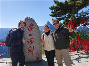 Three French tourists posing at Mount Hua with mountain peaks and red prayer ribbons in the background