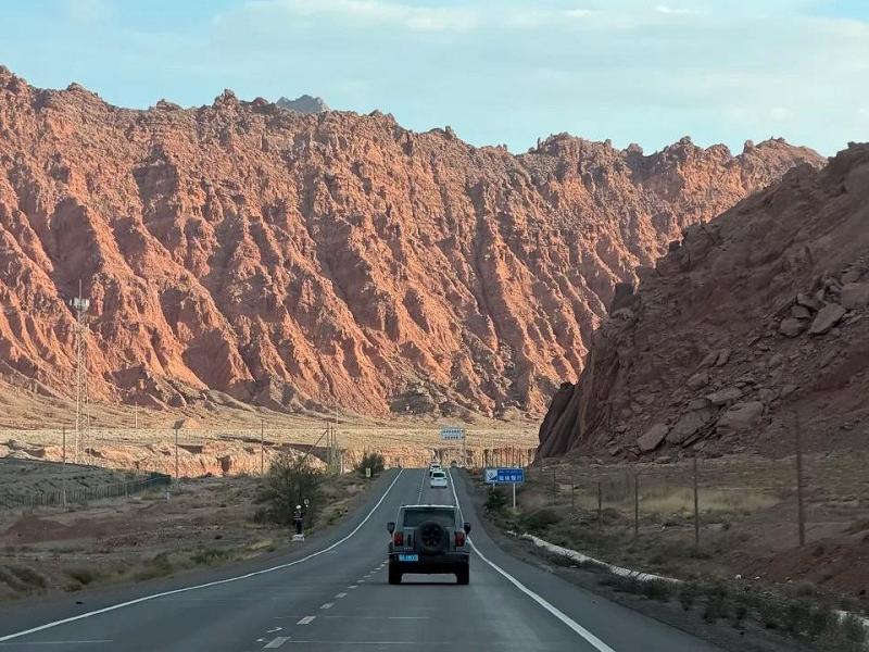 Scenic road through Xinjiang canyon with red rock mountains, desert landscape, and vehicles traveling under a blue sky