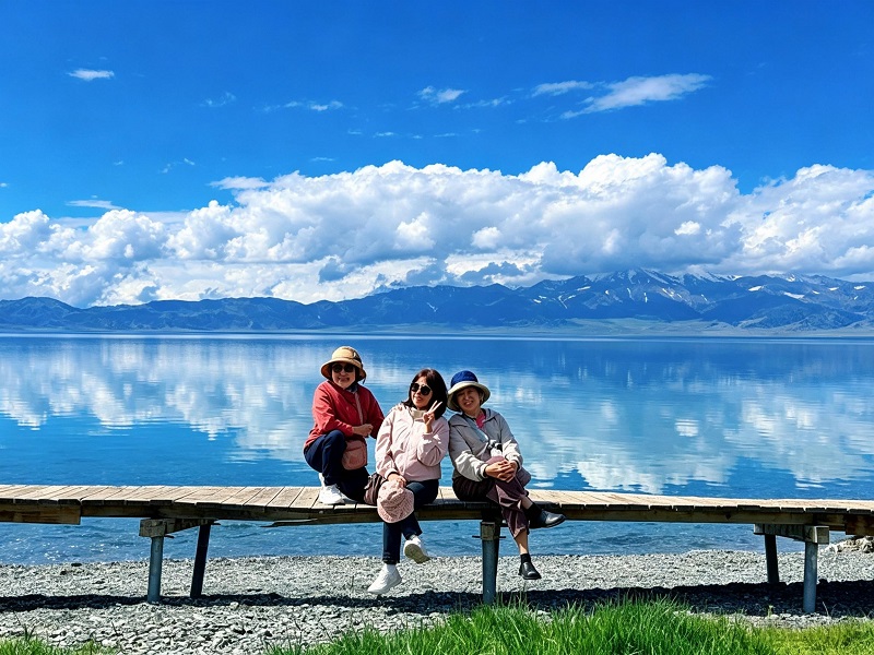 Three female travelers sitting on wooden pier, perfect cloud reflection on Sayram Lake with snow?capped Tianshan background
