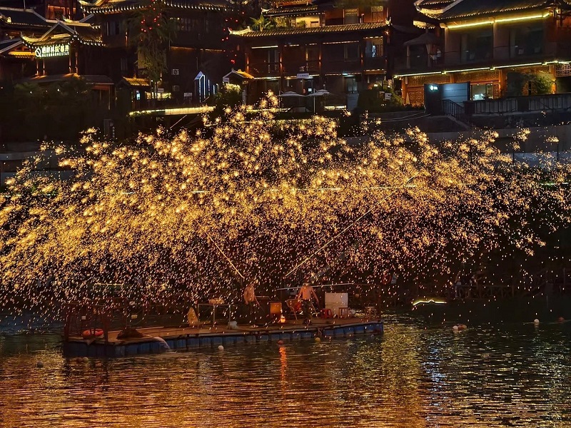 Traditional iron flower show on river floating stage, golden sparks reflecting over water, Dong-style buildings background