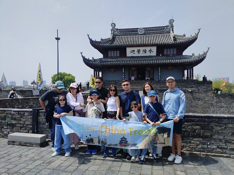 Tour group holding Buer China Travel banner, posing in front of Suzhou ancient city gate building on sunny day