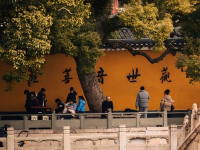 Bright orange temple wall carved with Chinese Guanyin calligraphy, ancient big tree and visiting pilgrims at Putuoshan Buddhist temple
